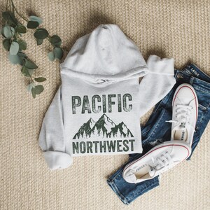 May include: A gray hooded sweatshirt with the text "PACIFIC NORTHWEST" in green with a mountain and tree graphic. The sweatshirt is laid out on a beige surface with a pair of white sneakers and blue jeans.