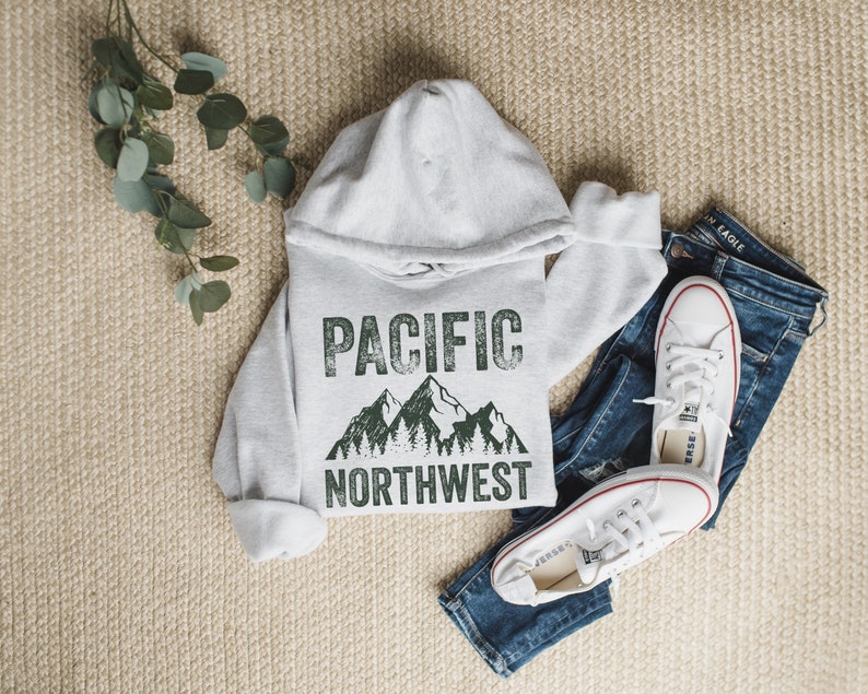 May include: A gray hooded sweatshirt with the text "PACIFIC NORTHWEST" in green with a mountain and tree graphic. The sweatshirt is laid out on a beige surface with a pair of white sneakers and blue jeans.
