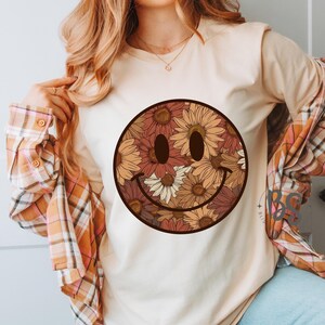 K&ouml;nnte beinhalten: Ein wei&szlig;es T-Shirt mit einem braunen und cremefarbenen floralen Smiley-Gesichtsdesign. Das Smiley-Gesicht besteht aus verschiedenen Blumen, darunter Sonnenblumen und G&auml;nsebl&uuml;mchen.