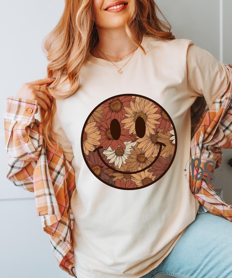 K&ouml;nnte beinhalten: Ein wei&szlig;es T-Shirt mit einem braunen und cremefarbenen floralen Smiley-Gesichtsdesign. Das Smiley-Gesicht besteht aus verschiedenen Blumen, darunter Sonnenblumen und G&auml;nsebl&uuml;mchen.