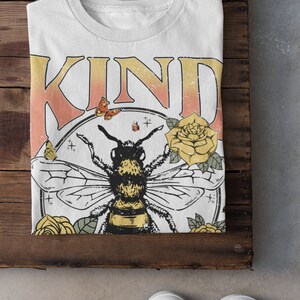 May include: White t-shirt with the word "KIND" in pink and orange letters. A bee with yellow and black stripes is surrounded by yellow flowers and butterflies in a circle.