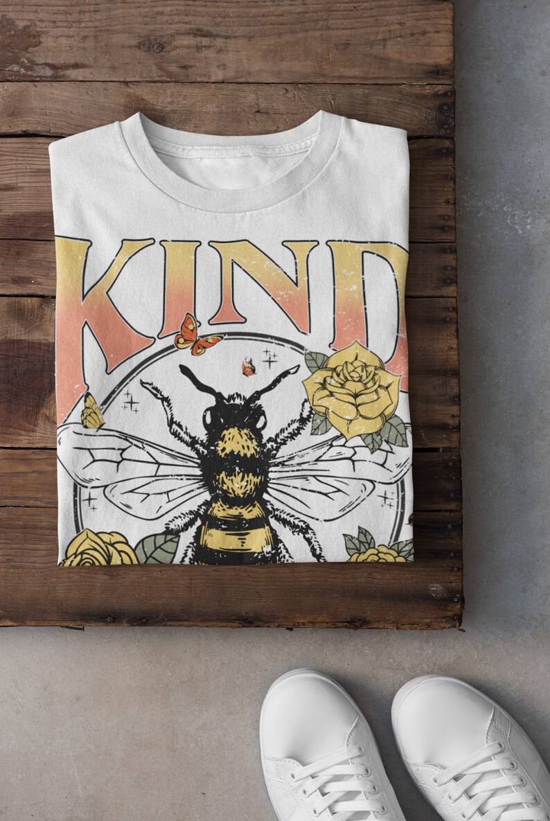 May include: White t-shirt with the word "KIND" in pink and orange letters. A bee with yellow and black stripes is surrounded by yellow flowers and butterflies in a circle.