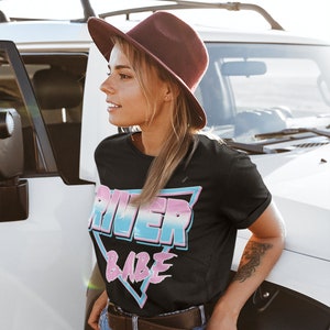 May include: A woman wearing a black t-shirt with a pink, blue, and white graphic that says "River Babe" in a retro style. She is wearing a brown hat and blue jeans.