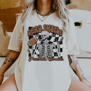 Puede incluir: Una camiseta blanca con un estampado gr&aacute;fico de un esqueleto sosteniendo un bal&oacute;n de f&uacute;tbol. El texto en la camiseta dice "Dead Inside But It's Football Season".