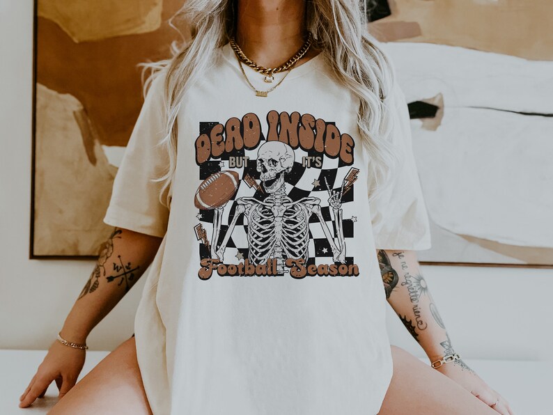 Puede incluir: Una camiseta blanca con un estampado gr&aacute;fico de un esqueleto sosteniendo un bal&oacute;n de f&uacute;tbol. El texto en la camiseta dice "Dead Inside But It's Football Season".