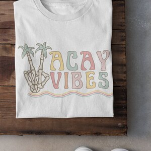May include: White t-shirt with a colorful graphic design that says "Vacay Vibes". The graphic features a skeleton hand making a peace sign with palm trees behind it.