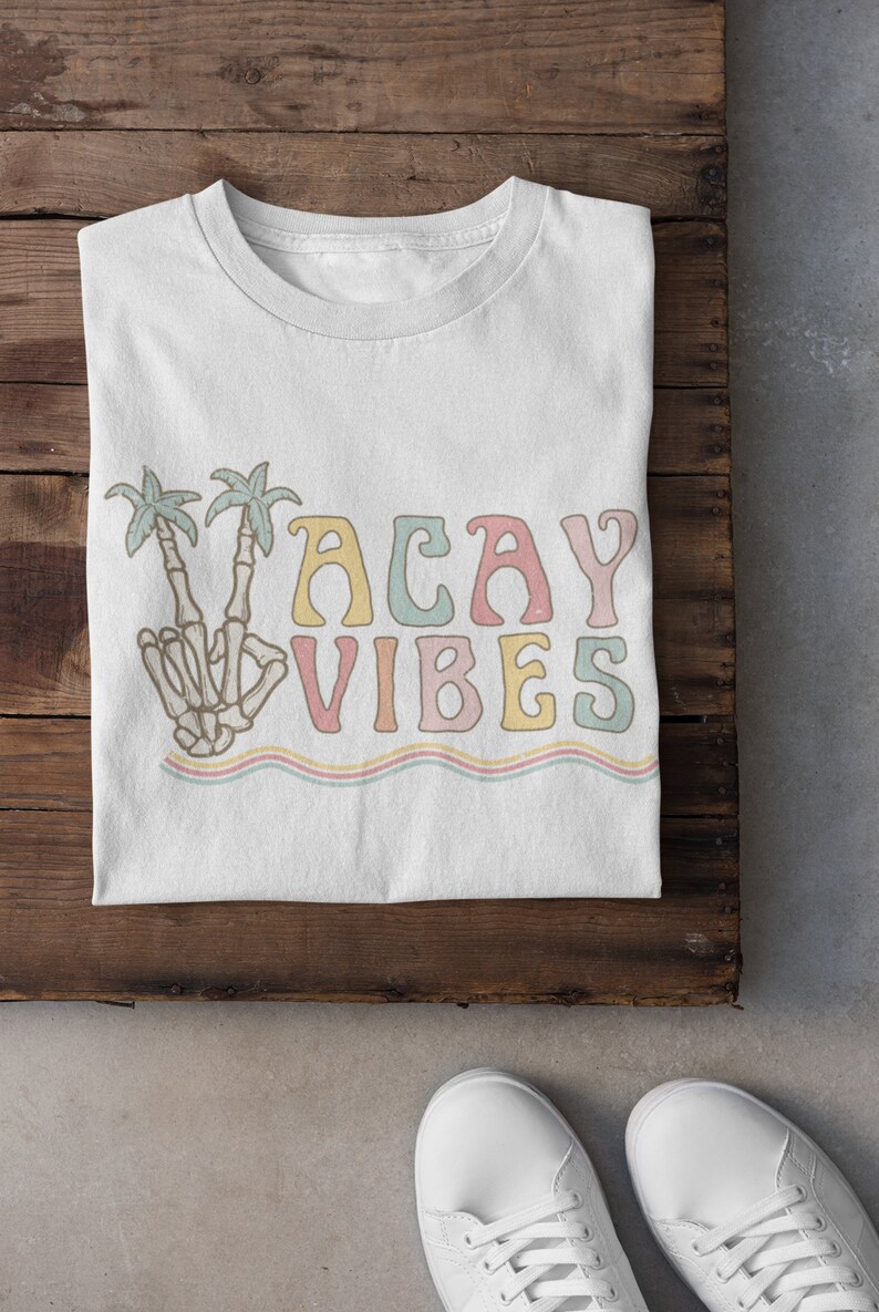 May include: White t-shirt with a colorful graphic design that says "Vacay Vibes". The graphic features a skeleton hand making a peace sign with palm trees behind it.