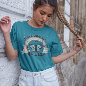 May include: Teal blue t-shirt with a rainbow, butterfly, and flowers graphic. The text on the shirt reads "Have Courage and Be Kind."
