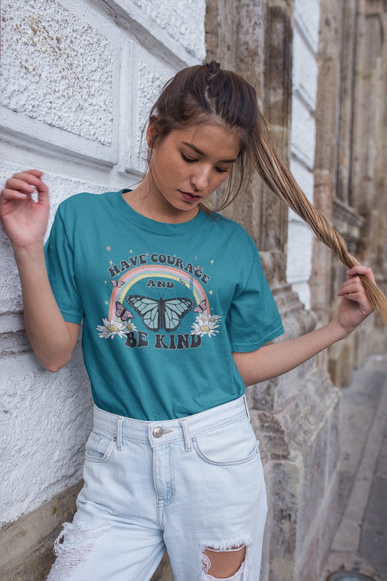 May include: Teal blue t-shirt with a rainbow, butterfly, and flowers graphic. The text on the shirt reads "Have Courage and Be Kind."