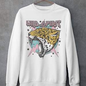 May include: White sweatshirt featuring a graphic print of a roaring jaguar head with a pink and blue background. The text "WILD SPIRIT" is above the jaguar head.