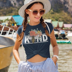 Pu&ograve; includere: Una maglietta blu navy con un design floreale e un'ape con la parola "KIND" stampata in bianco con un effetto vintage.