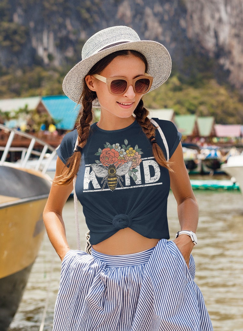 Pu&ograve; includere: Una maglietta blu navy con un design floreale e un'ape con la parola "KIND" stampata in bianco con un effetto vintage.
