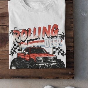 May include: White t-shirt with a red monster truck graphic and the text "Rolling Deep" in red and blue letters. The truck has a red body, black wheels, and a white cab. The background is a faded red, white, and blue design.