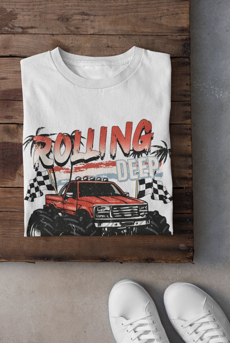 May include: White t-shirt with a red monster truck graphic and the text "Rolling Deep" in red and blue letters. The truck has a red body, black wheels, and a white cab. The background is a faded red, white, and blue design.