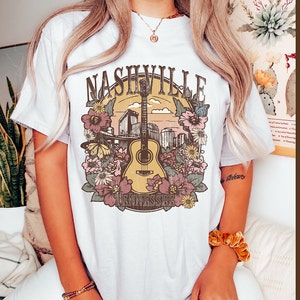 May include: White t-shirt with a graphic print of a guitar, flowers, and the text "Nashville Tennessee".