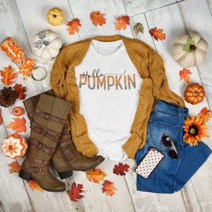 May include: A white t-shirt with the text "Hello Pumpkin" in orange and brown letters with a leopard print design. The shirt is paired with a brown cardigan, blue jeans, brown boots, and fall-themed decorations.
