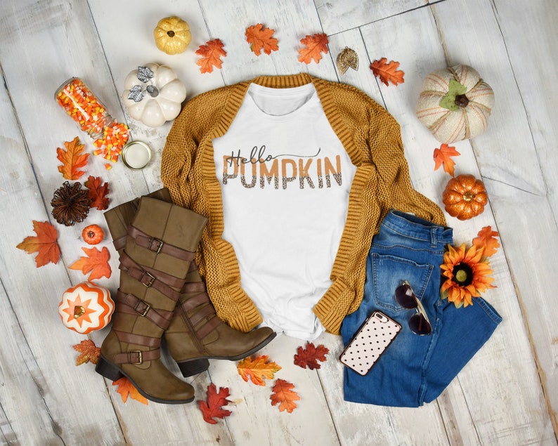May include: A white t-shirt with the text "Hello Pumpkin" in orange and brown letters with a leopard print design. The shirt is paired with a brown cardigan, blue jeans, brown boots, and fall-themed decorations.
