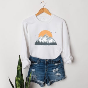 May include: White sweatshirt with a graphic of a mountain range with a yellow sun and blue trees.