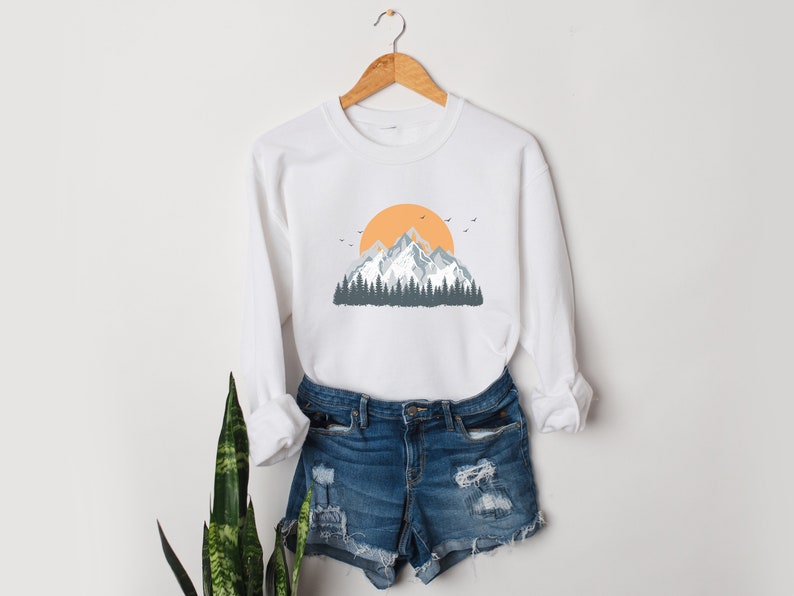May include: White sweatshirt with a graphic of a mountain range with a yellow sun and blue trees.