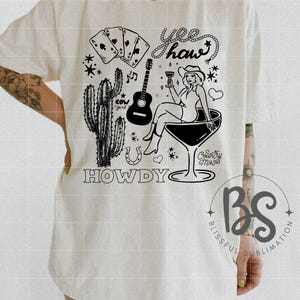 Pode incluir: Uma camiseta branca com um design gr&aacute;fico preto que apresenta uma vaqueira com um chap&eacute;u de vaqueiro, segurando uma ta&ccedil;a de vinho, sentada em uma ta&ccedil;a de martini. O design tamb&eacute;m inclui um cacto, uma ferradura, cartas de jogo, um viol&atilde;o, notas musicais e o texto "Yee haw" e "Howdy".