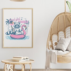 May include: A framed print with a cartoon raccoon wearing a life vest and holding a bottle of beer, floating on a pink flamingo pool float. The text "feral girl summer" is above the raccoon. The print is hanging on a white wall.