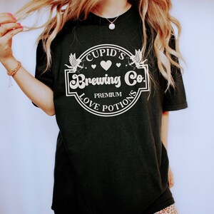 May include: Black t-shirt with a white graphic of a circle with the text "Cupid's Brewing Co. Premium Love Potions" and two white cherubs with arrows.