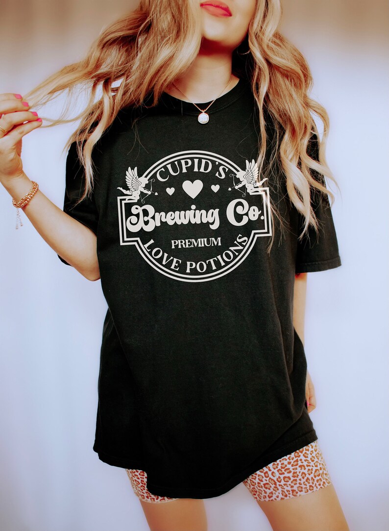 May include: Black t-shirt with a white graphic of a circle with the text "Cupid's Brewing Co. Premium Love Potions" and two white cherubs with arrows.