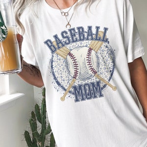 Op de afbeelding: Wit T-shirt met een blauwe en witte baseball afbeelding. De afbeelding toont een honkbal, twee gekruiste knuppels en de tekst "Baseball Mom".