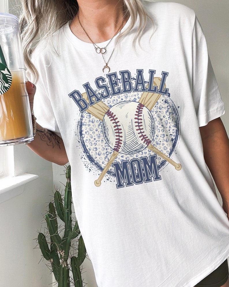 Op de afbeelding: Wit T-shirt met een blauwe en witte baseball afbeelding. De afbeelding toont een honkbal, twee gekruiste knuppels en de tekst "Baseball Mom".