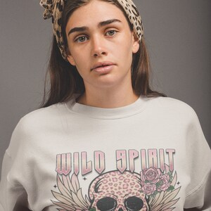 May include: A white sweatshirt with a pink and black skull graphic. The skull has a leopard print design and is surrounded by pink roses and wings. The text "WILD SPIRIT SWEET SOUL" is printed in pink on the sweatshirt.