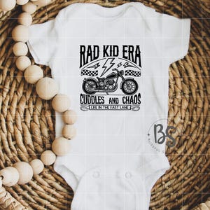 May include: White baby onesie with black text that reads "Rad Kid Era" with a lightning bolt and checkered flag design. Below the text is a black and white illustration of a motorcycle with the text "Cuddles and Chaos" and "Life in the Fast Lane".