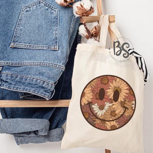 K&ouml;nnte beinhalten: Eine wei&szlig;e Canvas-Tragetasche mit einem braunen Smiley-Gesichtsdesign. Das Smiley-Gesicht ist mit einem Muster aus orangefarbenen, braunen und wei&szlig;en Blumen gef&uuml;llt. Die Tasche hat einen schwarz-wei&szlig; gestreiften Griff.