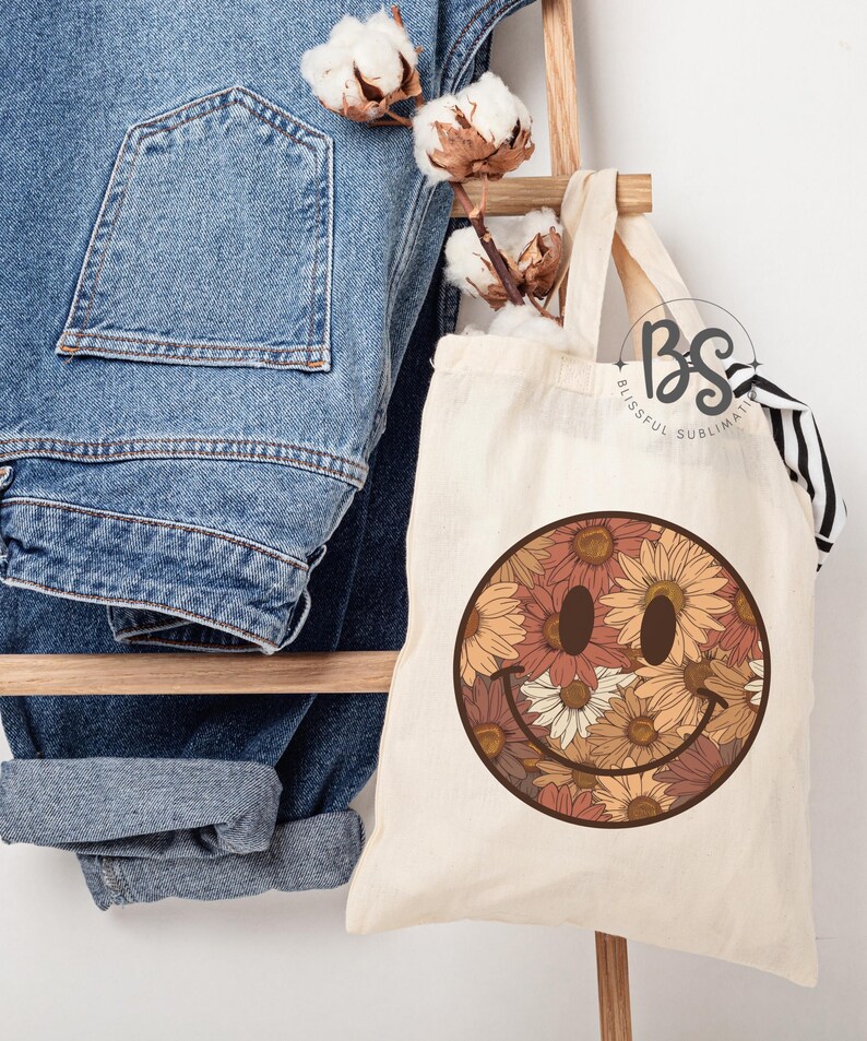 K&ouml;nnte beinhalten: Eine wei&szlig;e Canvas-Tragetasche mit einem braunen Smiley-Gesichtsdesign. Das Smiley-Gesicht ist mit einem Muster aus orangefarbenen, braunen und wei&szlig;en Blumen gef&uuml;llt. Die Tasche hat einen schwarz-wei&szlig; gestreiften Griff.