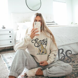 May include: A cream-colored sweatshirt with a graphic of a tiger with the word "Easy" in a stylized font. The tiger is yellow and black with a pink nose and tongue. The sweatshirt is worn by a person sitting on a patterned rug.