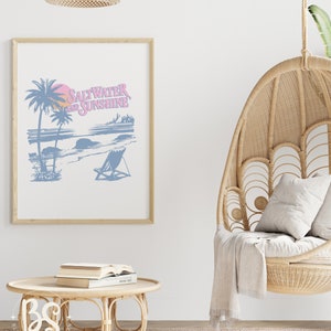 May include: A framed print with a blue and pink illustration of a beach scene with palm trees, a sunset, and a beach chair. The text "Saltwater and Sunshine" is written in pink on the print.