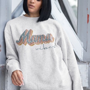 Puede incluir: Una sudadera gris con el texto "Mama Vibes" en una fuente retro arco&iacute;ris.