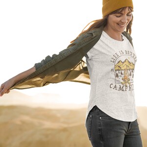 Pu&ograve; includere: Una donna indossa una maglietta grigia con la scritta "Life is better by the campfire" e una grafica con una scena di montagna. Indossa una camicia verde sopra la maglietta e un berretto giallo.