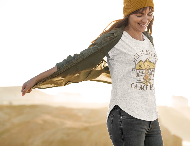 Pu&ograve; includere: Una donna indossa una maglietta grigia con la scritta "Life is better by the campfire" e una grafica con una scena di montagna. Indossa una camicia verde sopra la maglietta e un berretto giallo.