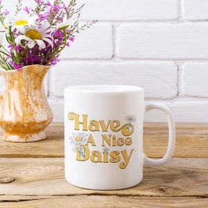 May include: White ceramic mug with a yellow and white daisy design and the text "Have A Nice Daisy". The mug is sitting on a wooden surface next to a vase with white and purple flowers.