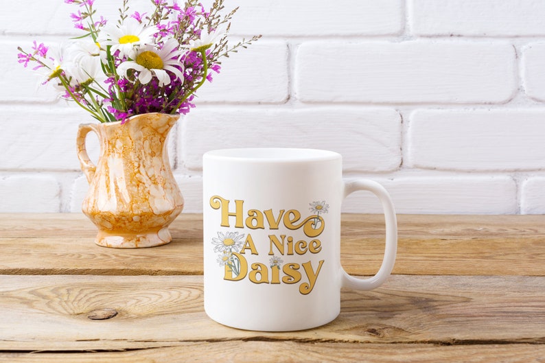 May include: White ceramic mug with a yellow and white daisy design and the text "Have A Nice Daisy". The mug is sitting on a wooden surface next to a vase with white and purple flowers.