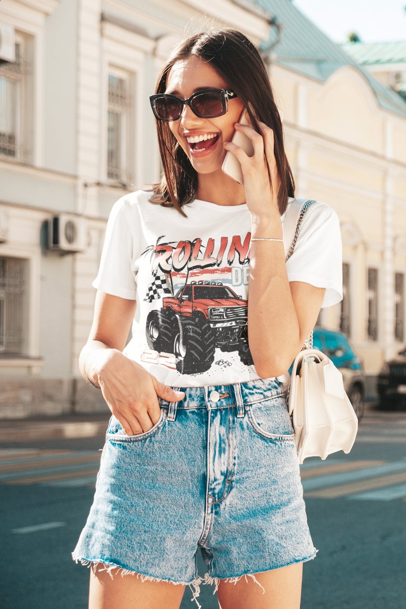 May include: A woman wearing a white t-shirt with a red, white, and blue graphic of a monster truck and the text "Rollin' Deep". She is also wearing blue denim shorts and sunglasses.