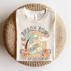 Pu&ograve; includere: Una t-shirt beige con un design grafico di una palma, onde oceaniche e un arcobaleno con il testo "Beach Bum Forever".