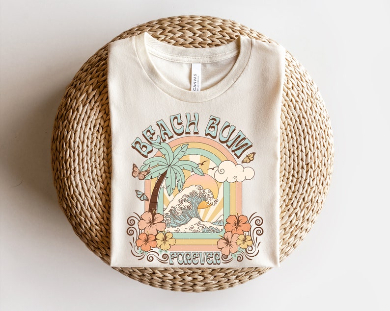 Pu&ograve; includere: Una t-shirt beige con un design grafico di una palma, onde oceaniche e un arcobaleno con il testo "Beach Bum Forever".