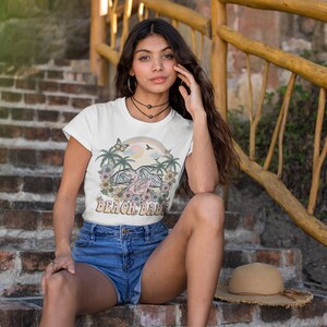 Op de afbeelding: Een wit T-shirt met een kleurrijke afbeelding van palmbomen, een regenboog en de tekst "Beach Babe". Het model draagt het shirt met een spijkerbroekje en zit op een trap van bakstenen.