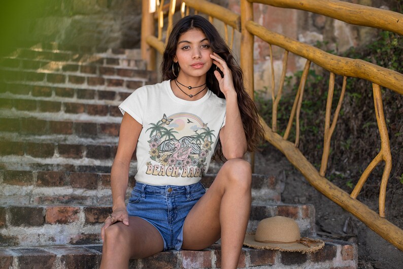 Op de afbeelding: Een wit T-shirt met een kleurrijke afbeelding van palmbomen, een regenboog en de tekst "Beach Babe". Het model draagt het shirt met een spijkerbroekje en zit op een trap van bakstenen.