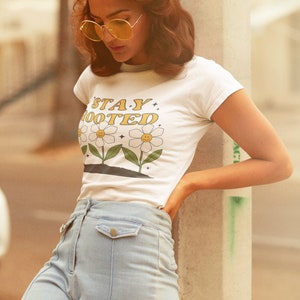 May include: A woman wearing a white t-shirt with the text "STAY ROOTED" and a daisy graphic. She is also wearing light blue jeans and yellow sunglasses.