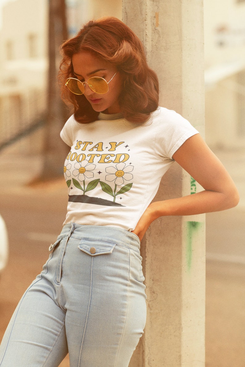 May include: A woman wearing a white t-shirt with the text "STAY ROOTED" and a daisy graphic. She is also wearing light blue jeans and yellow sunglasses.