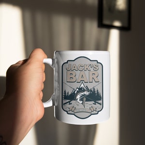 May include: White ceramic mug with a brown and gray graphic of a fish jumping out of the water with fishing rods and the text "Jack's Bar Virgin River, CA."