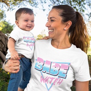 May include: A white t-shirt with the text "Babe Watch" in a retro, colorful design. A child wearing a white t-shirt with the text "Little Babe" in a similar design is being held by the person wearing the "Babe Watch" shirt.