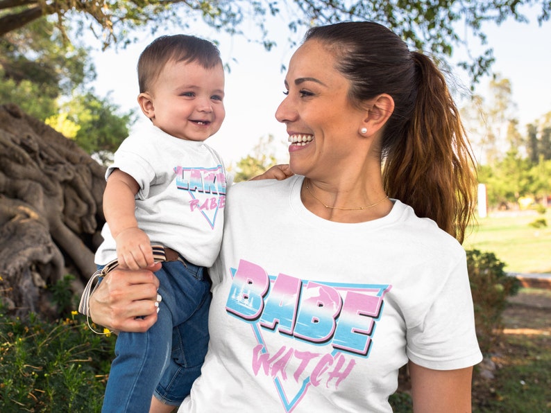 May include: A white t-shirt with the text "Babe Watch" in a retro, colorful design. A child wearing a white t-shirt with the text "Little Babe" in a similar design is being held by the person wearing the "Babe Watch" shirt.